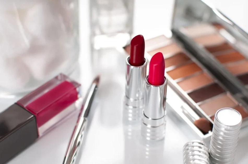 A vibrant flat-lay arrangement of foundation bottles, lip tints, and makeup products in unmarked packaging placed on a clean white and gold vanity surface, representing the makeup category portion of the luxury versus drugstore blind comparison test conducted by InfoProds in 2026