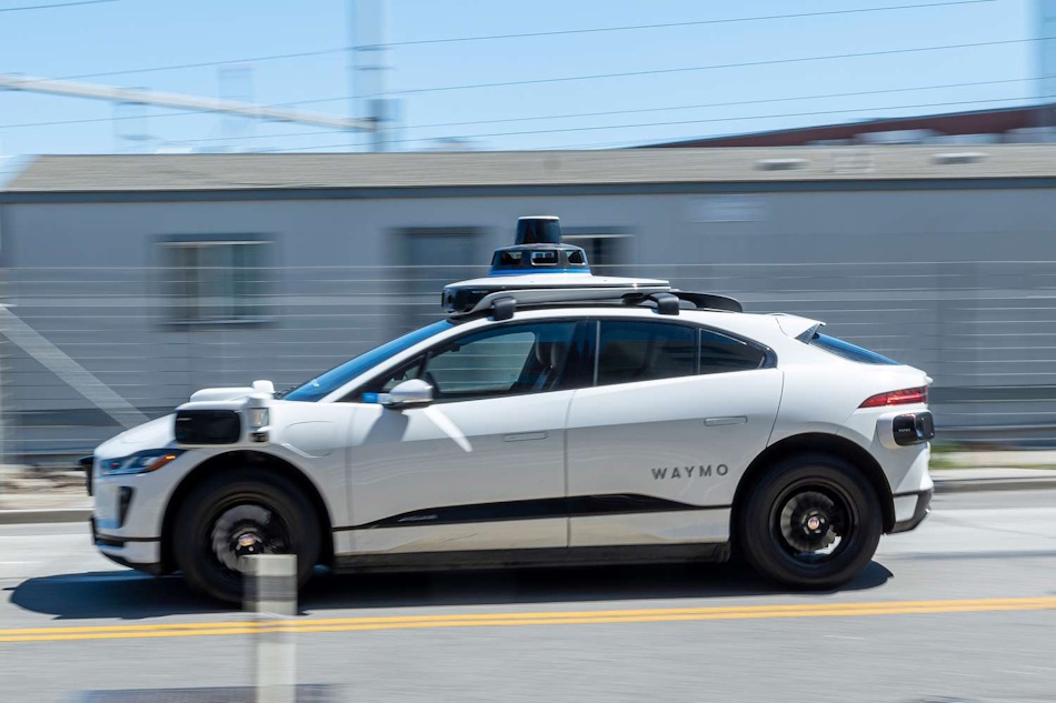 White Waymo autonomous robotaxi with distinctive sensor dome navigating through busy San Francisco street intersection with pedestrians and vehicles, showcasing advanced self-driving technology and urban mobility solutions in 2026 - InfoProds automotive technology guide