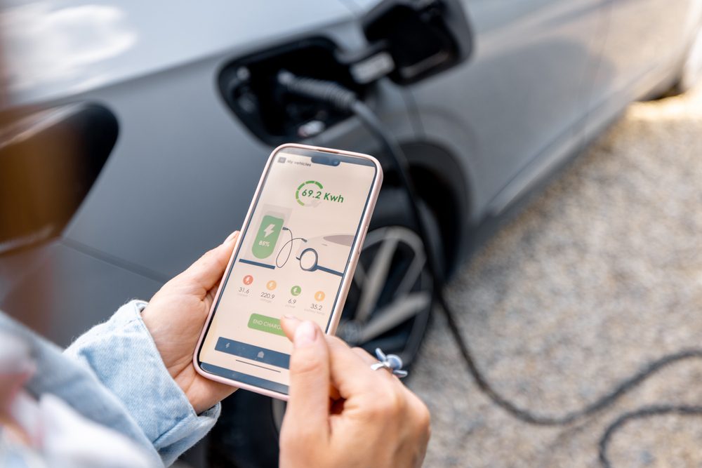 Electric vehicle owner using smartphone app to manage charging schedule and electricity costs EV owner using mobile charging app to schedule off-peak charging optimize time-of-use electricity rates and manage total ownership costs - InfoProds 2026