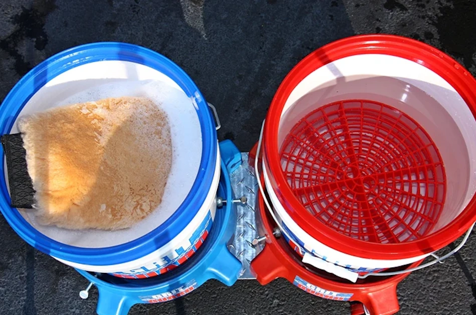 Proper two-bucket car washing setup featuring separate wash and rinse buckets with grit guards at bottom, premium microfiber wash mitt, pH-neutral car wash soap, and foam cannon attachment preventing swirl marks during vehicle cleaning through systematic dirt management and paint-safe technique - InfoProds 2026