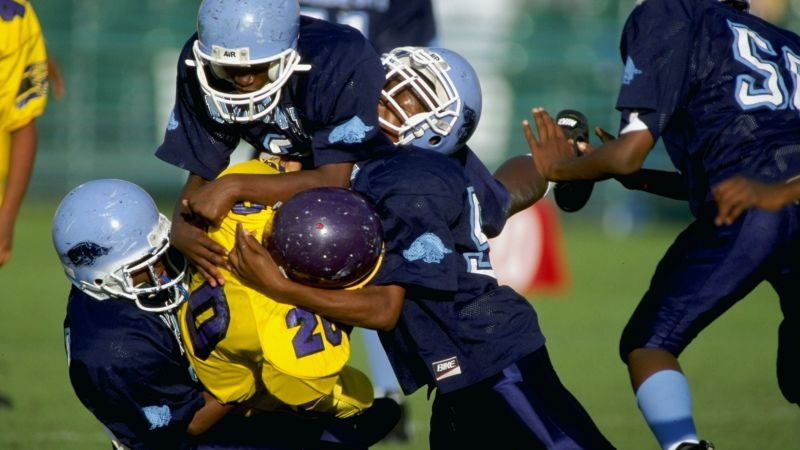 Youth Football Practice Showing Young Players Head Impact Exposure Risk Young children participating in tackle football practice demonstrating early age exposure to repetitive head impacts that research links to increased chronic traumatic encephalopathy risk, with studies showing youth tackle players sustain 15 times more head impacts than flag football participants during equivalent playing time - InfoProds Youth Sports Safety Research 2026