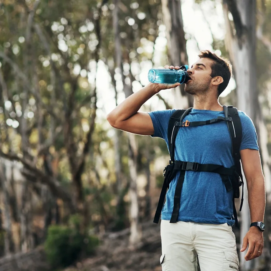 Hiker resting in shade drinking water during summer trail trek Exhausted hiker sitting in natural shade taking hydration break with water bottle during summer mountain trail hike, demonstrating heat illness prevention strategies and proper rest techniques to avoid heatstroke during outdoor adventure activities - InfoProds 2026