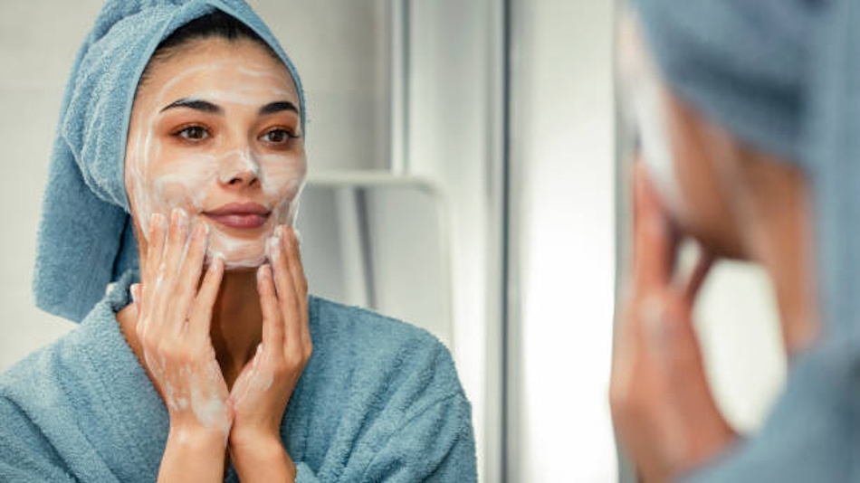 Young woman in modern bathroom examining her facial skin closely in mirror surrounded by extensive collection of skincare serums bottles toners and treatment products scattered across counter representing overwhelming complexity of modern beauty routines before discovering dermatologist-approved skin cycling method - InfoProds Beauty Guide 2026