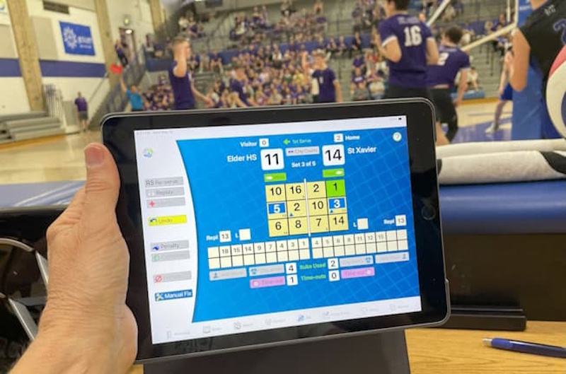 A coach’s hand holding a high-tech tablet displaying a 3D volleyball rotation chart and player positioning data during a 2026 match - InfoProds 2026